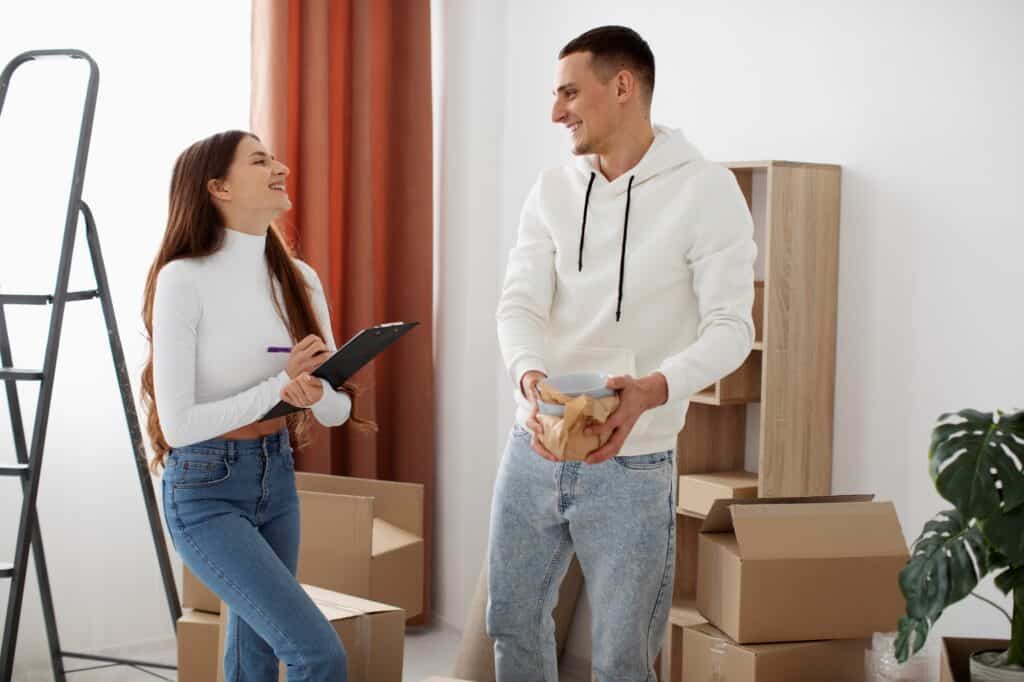 A couple surrounded by boxes and ladder, holding the checklist in the concept of 'how to plan and prepare for home removals in East London'.