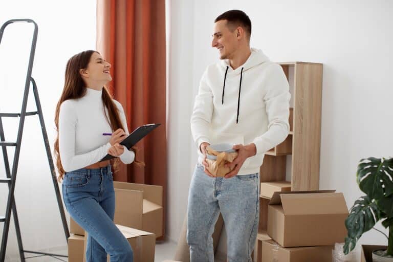 A couple surrounded by boxes and ladder, holding the checklist in the concept of 'how to plan and prepare for home removals in East London'.